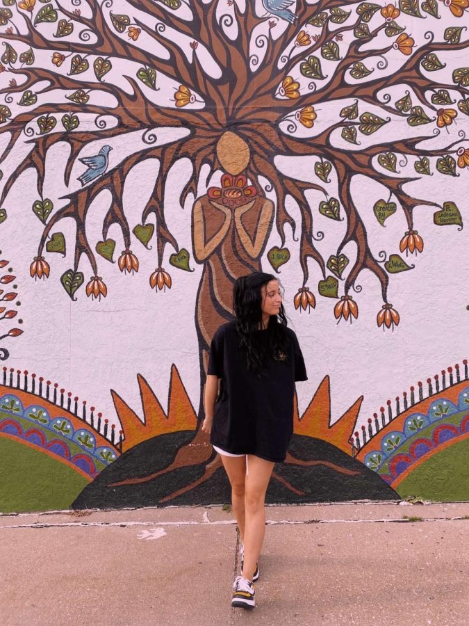 Riley Brandt walking in front of a creative mural representing a tree of life. Brandt has emphasized to others the positive changes she has seen in her life from practicing self-care. 
