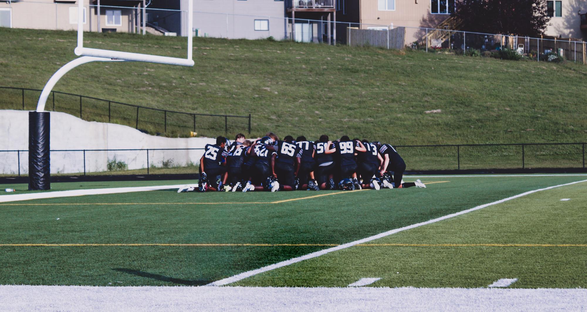 The freshman football team is crouched in a huddle before the game. This is their first home game of the season and they kneeled together to talk.
